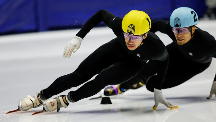 Un patineur de vitesse, vêtu d'une combinaison noire et d'un casque jaune, se penche pour effectuer un virage. 