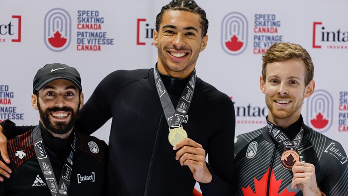 Trois hommes, médaille autour du cou, prennent la pose sur un podium après une compétition de patinage de vitesse sur courte piste. 