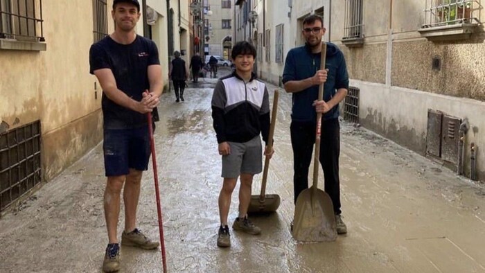 Trois hommes posent pour la photo avec des pelles et des balais dans une rue pleine de boue. 