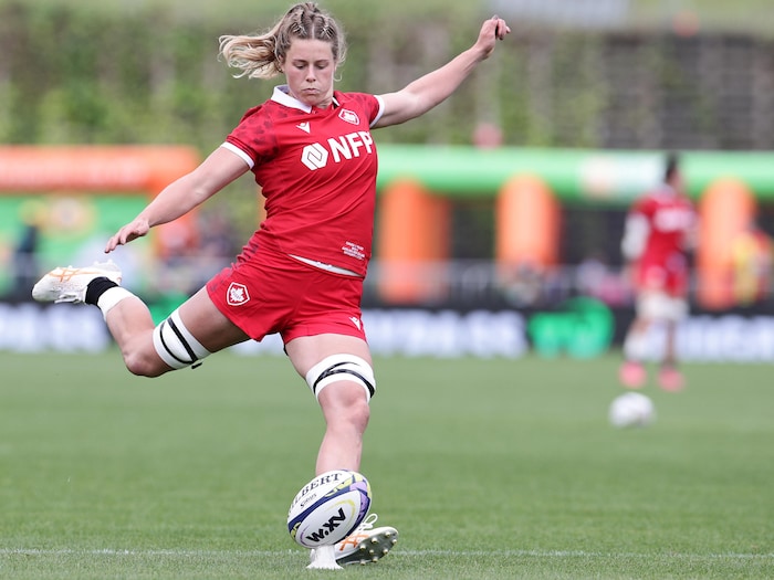 A rugby player is in full swing, arms outstretched, about to kick the ball with her right foot.