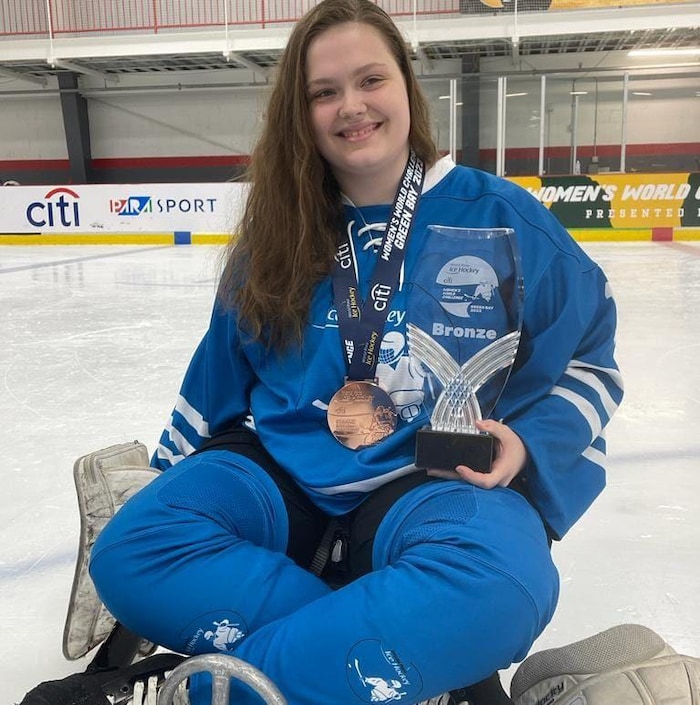Elle tient un trophée et porte une médaille de bronze autour du cou.