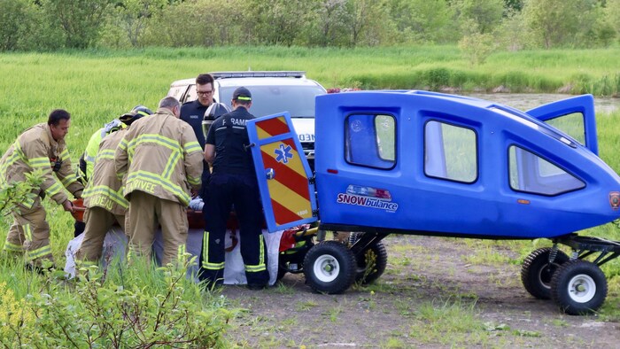 Des pompiers et des policiers entourent la civière en la sortant de la remorque fermée.