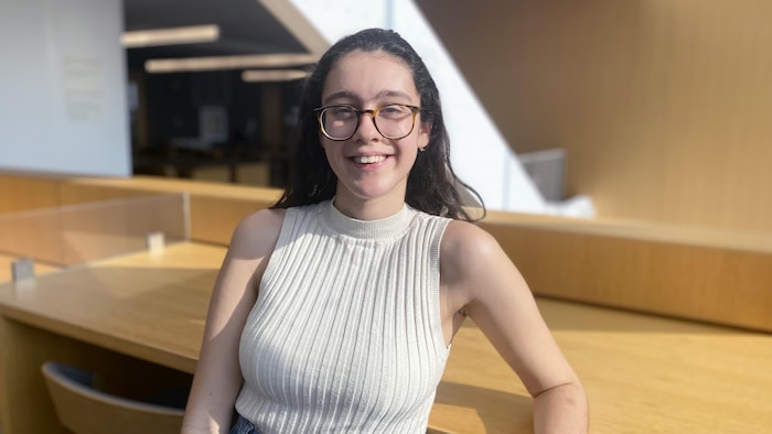 Une femme (Maya Larrondo, petite-fille d'exilés chiliens au Canada et étudiante en anthropologie médico-légale à l'université de Toronto) sourit à la caméra dans une bibliothèque. 