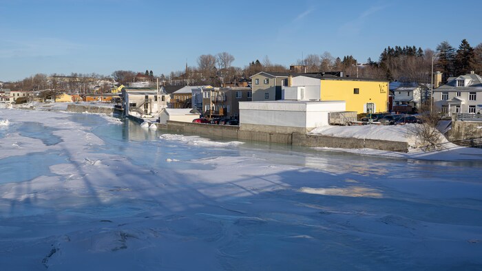 Un mur de béton longe une rivière. Le dos de nombreux bâtiments se trouvent sur cette berge.