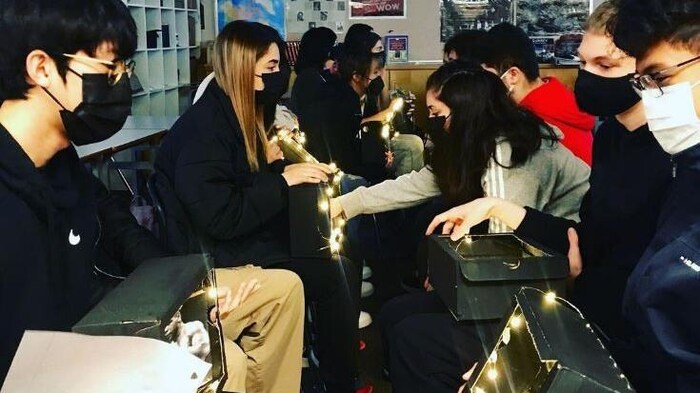 Une rangée d'étudiants masqués sont assis avec des boîtes lumineuses sur leurs genoux, en face d'une autre rangée d'étudiants masqués dans une salle de classe.