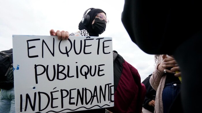 Une manifestante portant un masque tient une pancarte où l'on peut lire «Enquête publique indépendante».