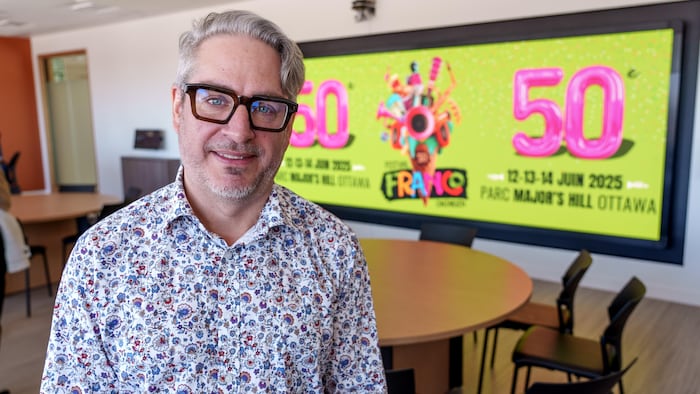 José Bertrand sourit à la caméra. En arrière-plan, une affiche officielle du Festival franco-ontarien.