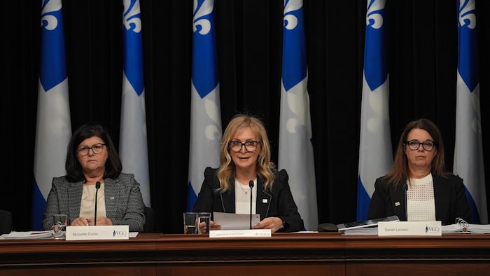 Janique Lambert entourée de Moïsette Fortin et de Sarah Leclerc en conférence de presse devant des drapeaux du Québec.