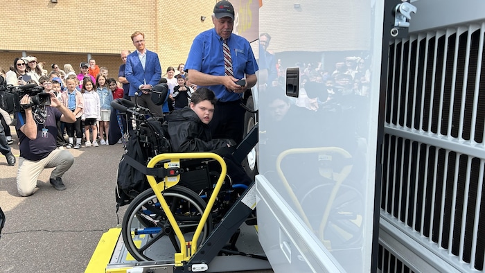 Un jeune garçon en fauteuil roulant sur une rampe, s'apprêtant à monter à bord d'un autocar, un chauffeur et une foule derrière lui.