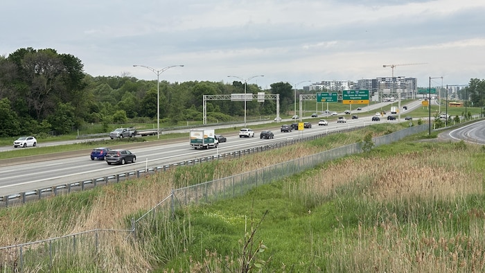 Vue large de l'autoroute Robert-Bourassa, une voie rapide à plusieurs voies avec des véhicules circulant dans les deux sens sous un ciel partiellement nuageux. Des panneaux de signalisation routière sont visibles au-dessus des voies. De l'herbe haute et des roseaux bordent les accotements de l'autoroute au premier plan, tandis qu'une dense zone boisée s'étend à gauche.