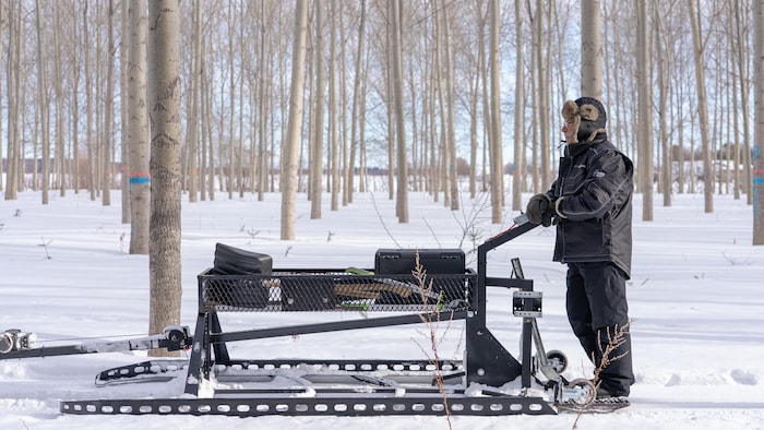 Un homme habillé chaudement se tient debout sur la plateforme d'un dispositif de traçage de piste de ski de fond, dans une plantation enneigée.