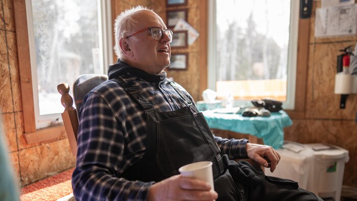 Robert Lavoie est assis dans une chaise berçante dans un chalet, un gobelet de café à la main.