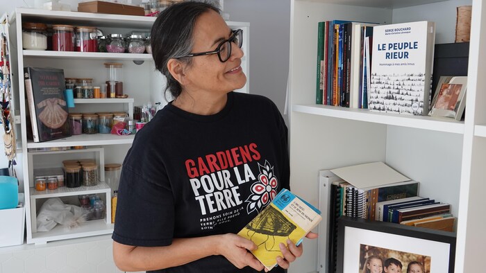 Michèle Audette regarde des livres dans sa bibliothèque et tient dans sa main le livre « Carcajou et le sens du monde ».