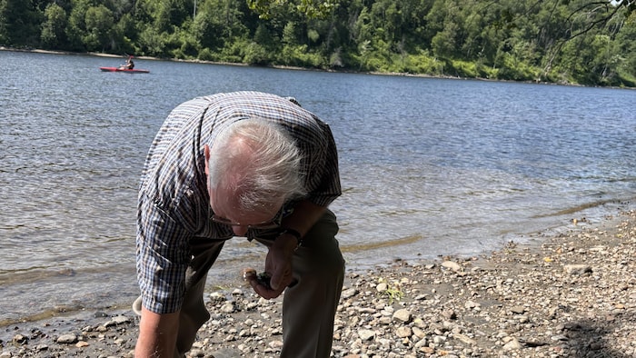 Marcel Pelchat est penché pour ramasser des morceaux de verre qui traînent sur la Plage à chien. Derrière lui, la rivière Saint-Maurice.