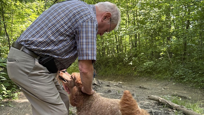 Marcel Pelchat de profil, à gauche de l’image, joue avec un caniche haut sur pattes, à sa droite, dans un sentier menant à la Plage à chiens.