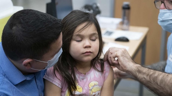 Une petite fille se fait vacciner, les yeux fermés. Son père, masqué, la réconforte.