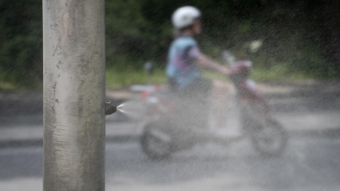 Une personne sur un scooter passe derrière un poteau qui diffuse de l'eau pendant une canicule.