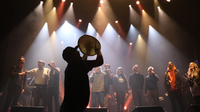 Un homme dans l'ombre avec un tambour et des chanteurs derrière.