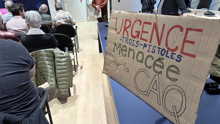 Une affiche militante indiquant urgence Trois-Pistoles menacée par la CAQ posée sur une table dans une salle remplie d'une centaines de personnes.