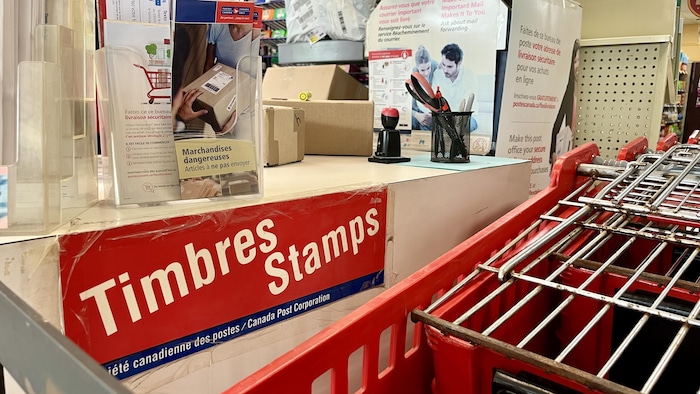 Une affiche qui porte le mot timbres est collée sur un comptoir postal où trônent des colis et des dépliants.