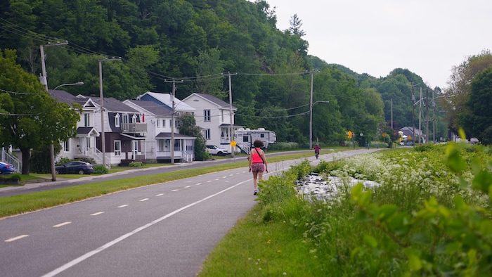 Une personne marche et une autre fait du vélo sur une piste cyclable pavée qui s'étend vers l'horizon. Sur la gauche, une rangée de maisons résidentielles avec des balcons et des façades de couleurs claires est visible. À l'arrière-plan et sur la droite, des collines densément boisées et des lignes électriques s'étendent. Le côté droit du chemin est bordé d'une végétation dense avec des fleurs blanches, et un petit cours d'eau serpente le long du chemin.