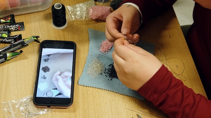 Des mains qui tiennent de petites perles au-dessus d'une table sur laquelle est déposé un téléphone intelligent qui diffuse une vidéo.