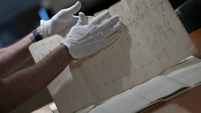 Des mains gantées tournent les pages d'un fragile document d'époque.