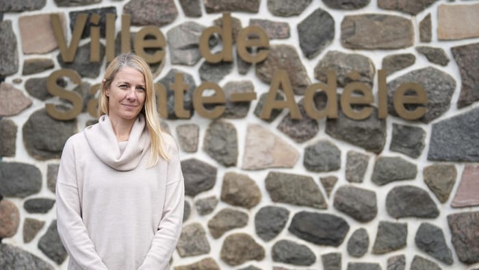 La mairesse se tient devant un mur de pierres sur lequel est écrit « Sainte-Adèle ».