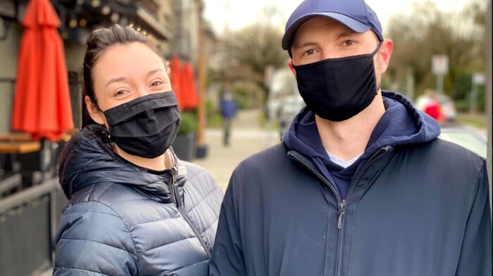 Emilie Roy et Robert Bryce, portant un masque et des vêtements d'automne, sont photographiés sur un trottoir, près d'une terrasse.