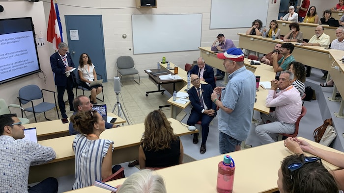 Une salle de classe dans laquelle des politiciens et des chercheurs discutent. La salle écoute un homme qui porte un chapeau aux couleurs de l'Acadie.