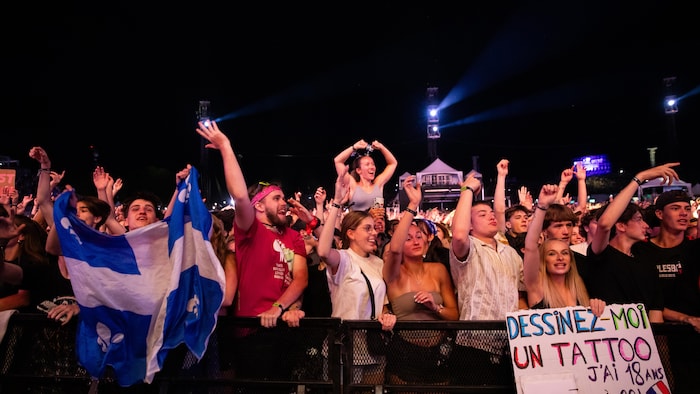 Un groupe de jeunes adultes est debout devant une barrière métallique lors d'un concert en plein air la nuit. Plusieurs lèvent les bras en l'air, certains ont des drapeaux ou des pancartes. Au centre droit, une pancarte manuscrite indique "DESSINEZ-MOI UN TATTOO J'AI 18 ANS À 00H".