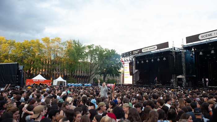 Une vaste foule de personnes est rassemblée en plein air pour un concert de musique. L'image est prise depuis le milieu de la foule, regardant vers la scène. Au premier plan, de nombreux spectateurs sont visibles, la plupart tournant le dos à l'appareil photo, absorbés par la scène.