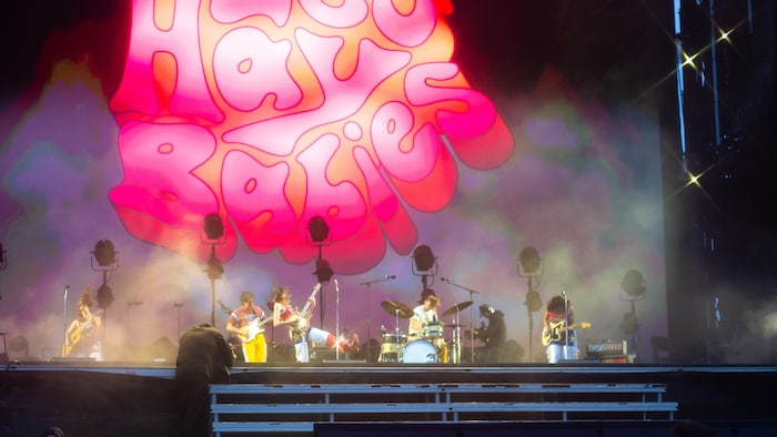 Une scène de concert vibrante avec un grand écran en arrière-plan affichant le nom du groupe "Les Hay Babies" en lettres roses et rouges de style psychédélique, entourées de brume. Plusieurs musiciens sont visibles sur scène. 