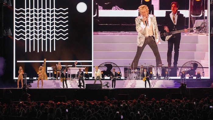 Image d'un concert en plein air montrant Rod Stewart sur scène, entouré de ses musiciens et de danseuses. Il porte une veste scintillante et un pantalon rayé, chantant avec énergie au micro.
