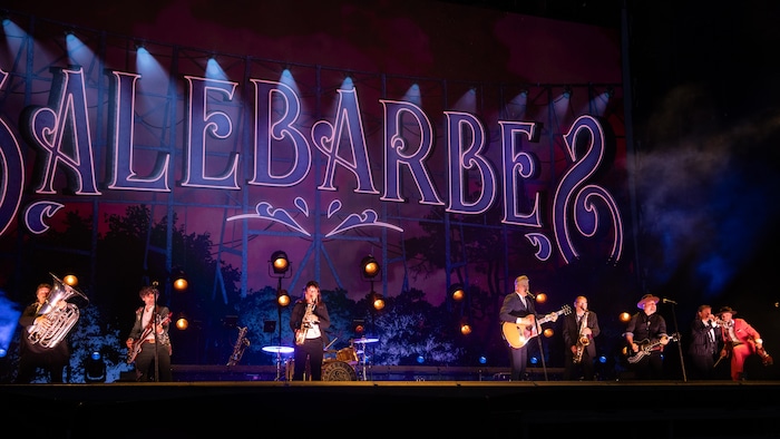 Photographie pleine largeur du groupe "Salebarbes" en concert sur une scène extérieure sombre, illuminée par des lumières vives roses et bleues. Le nom du groupe, "SALEBARBES", est affiché en grandes lettres fantaisistes lumineuses à l'arrière-plan. Sept musiciens sont visibles, chacun avec leur instrument, dont un sousaphone, plusieurs guitares électriques et acoustiques, une batterie et un saxophone. 