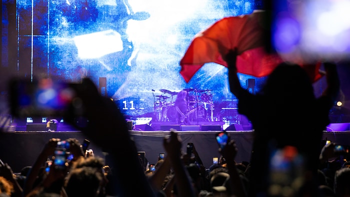 Une foule de spectateurs, dont certains tiennent des téléphones portables pour filmer, assiste à un concert nocturne. Au centre, sur une scène illuminée par des lumières bleues et blanches. Une personne au premier plan brandit un drapeau rouge.