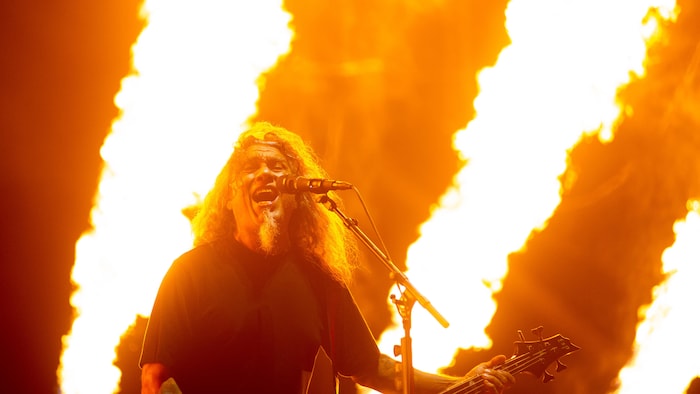 Tom Araya joue de la basse en chantant devant des flammes.