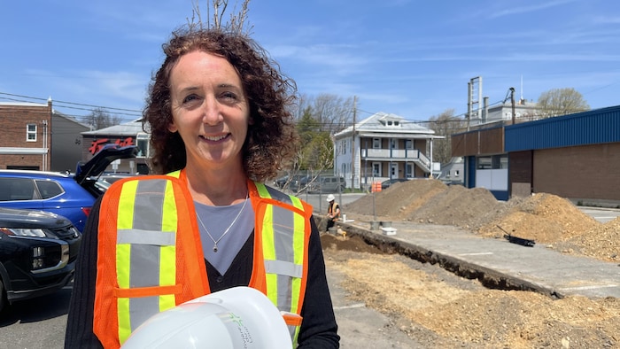 Julie Martin avec une veste de sécurité et un casque de construction devant une tranchée dans laquelle des archéologues creusent et s'affairent.