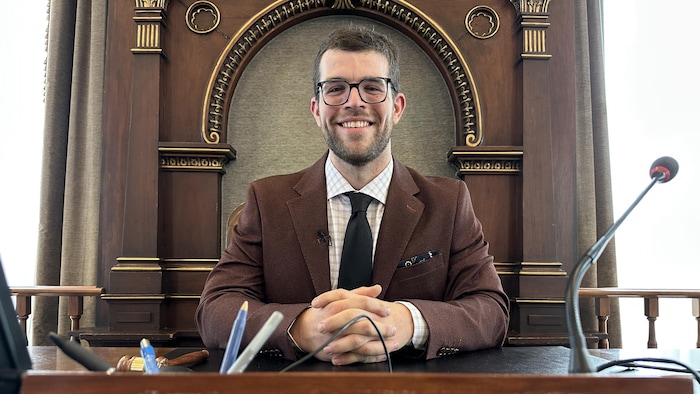 Un homme portant des lunettes siège derrière un bureau en bois.
