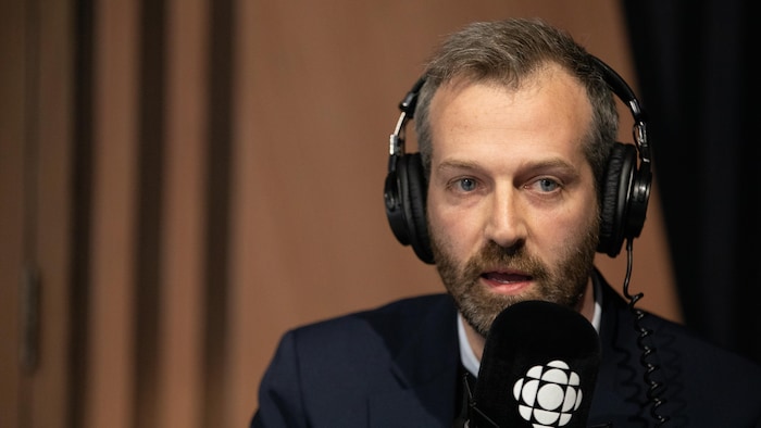 Un homme barbu aux cheveux bruns, portant un costume sombre et des écouteurs noirs, parle dans un microphone noir sur lequel est visible le logo de Radio-Canada. Son regard est dirigé vers l'avant, et l'arrière-plan est flou, avec des tons de bois.