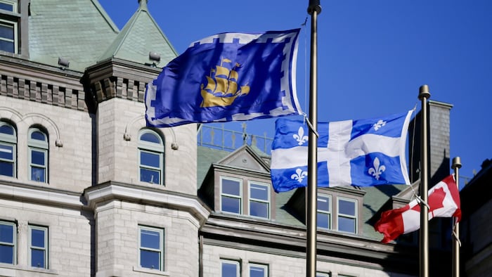 Des drapeaux de la Ville de Québec, du Québec et du Canada flottent au vent devant l'hôtel de ville de Québec.