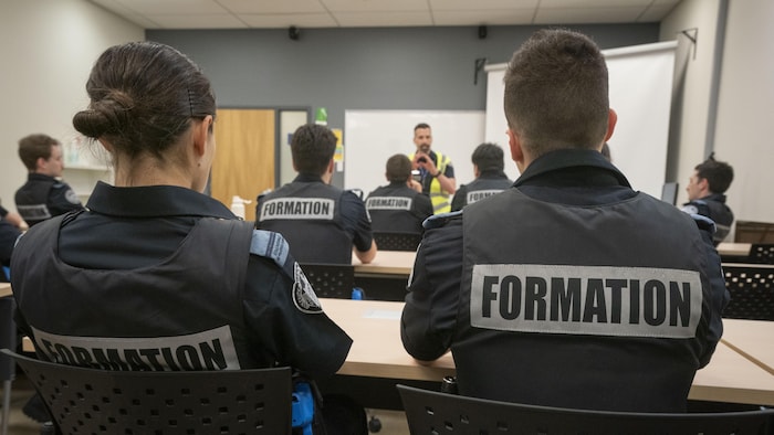 Des personnes portant un uniforme policier marqué de l'inscription « Formation », vues de dos, sont assises dans une salle de cours et écoutent un instructeur.