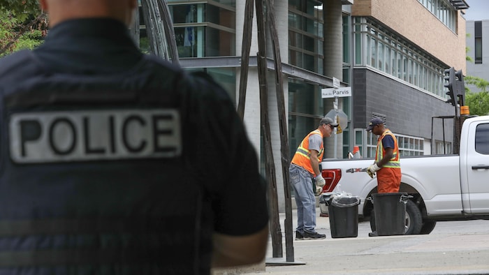 Un policier surveille deux employés municipaux faisant le ménage sur la rue du Parvis.