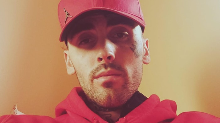 Un homme portant une casquette et un chandail rouge regarde la caméra. On voit qu'il a des tatouages sur le visage et le cou.