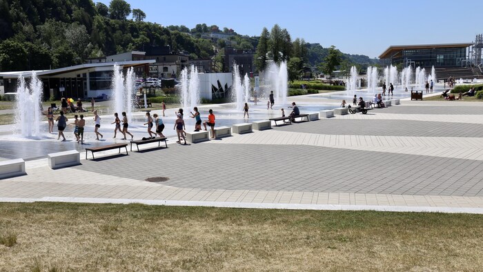 Vue grand-angle d'une place publique animée avec des jeux d'eau, le Quai Paquet à Lévis. Plusieurs jets d'eau verticaux créent des fontaines où de nombreux enfants et quelques adultes s'amusent.