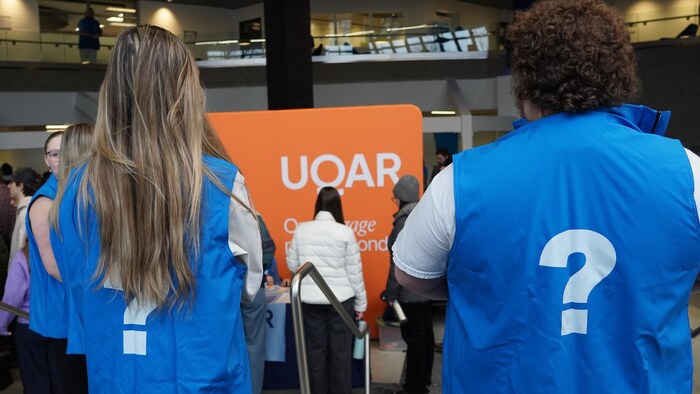 Deux jeunes personnes sont de dos et ont un point d'interrogation sur leur veste. Elles regardent un panneau qui porte la mention « UQAR ».
