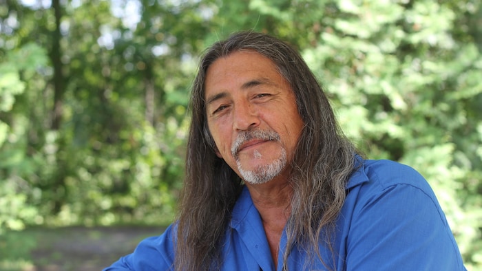 Serge Otsi Simon se tient souriant devant des arbres.