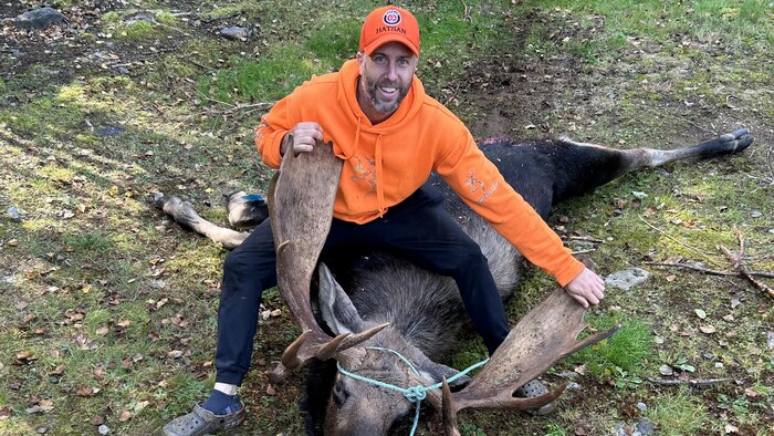 Remi Arseneault souriant, assis sur un orignal mort et tenant les cornes de bois de l'animal.