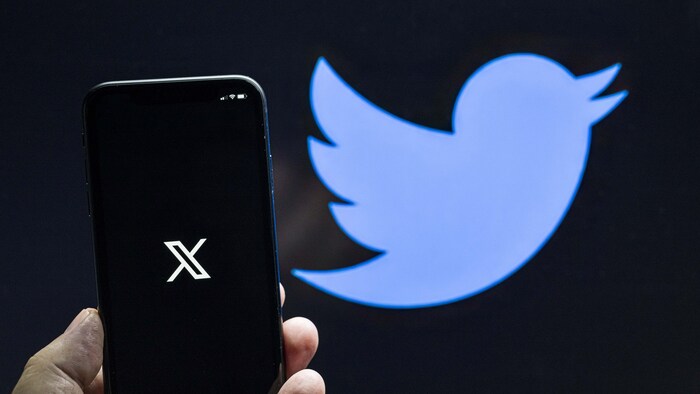 Un main tient un téléphone avec le logo de X devant un mur avec l'ancien logo de Twitter.