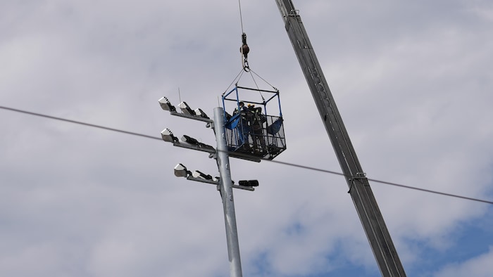 Des travailleurs sont dans une plateforme suspendue par une grue, près d'un poteau de plusieurs lumières.